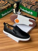 Black Timberland leather slip-on sneakers with white midsole and brown tread displayed with Timberland boxes and gold decor.