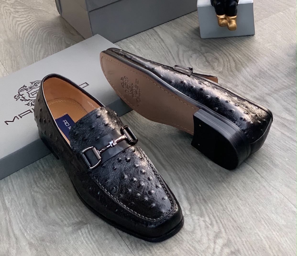 Genuine ostrich leather horsebit loafers Men's brown genuine ostrich leather horsebit loafers with square toe and rubber sole, displayed on grey wood floor with branded shoebox.
