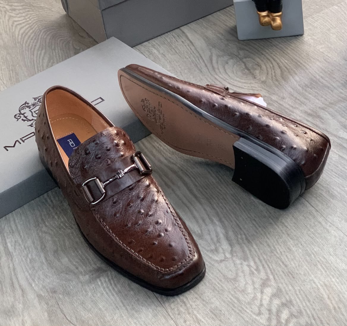 Genuine ostrich leather horsebit loafers Men's brown genuine ostrich leather horsebit loafers with square toe and rubber sole, displayed on grey wood floor with branded shoebox.
