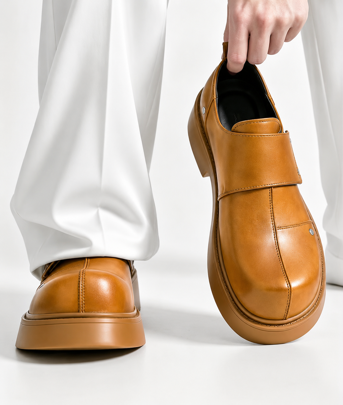 Premium leather monk-strap loafers Men's luxury leather monk-strap loafers with chunky black platform soles and silver hardware accents being held by hand next to white trousers.