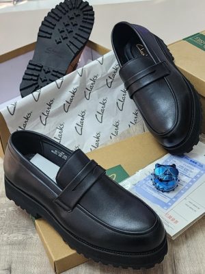 A pair of men's luxury tan leather loafers with a black chunky lug sole, displayed on a cardboard box with branded tissue paper, showcasing their sleek design and premium material.