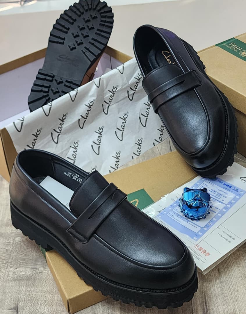 Men’s Luxury Leather Loafers - Clarks. A pair of men's luxury tan leather loafers with a black chunky lug sole, displayed on a cardboard box with branded tissue paper, showcasing their sleek design and premium material.