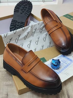 A pair of men's luxury tan leather loafers with a black chunky lug sole, displayed on a cardboard box with branded tissue paper, showcasing their sleek design and premium material.