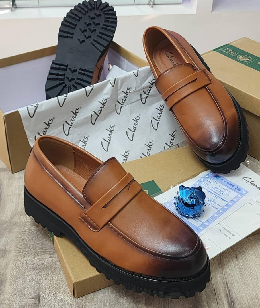 Men’s Luxury Leather Loafers - Clarks. A pair of men's luxury tan leather loafers with a black chunky lug sole, displayed on a cardboard box with branded tissue paper, showcasing their sleek design and premium material.