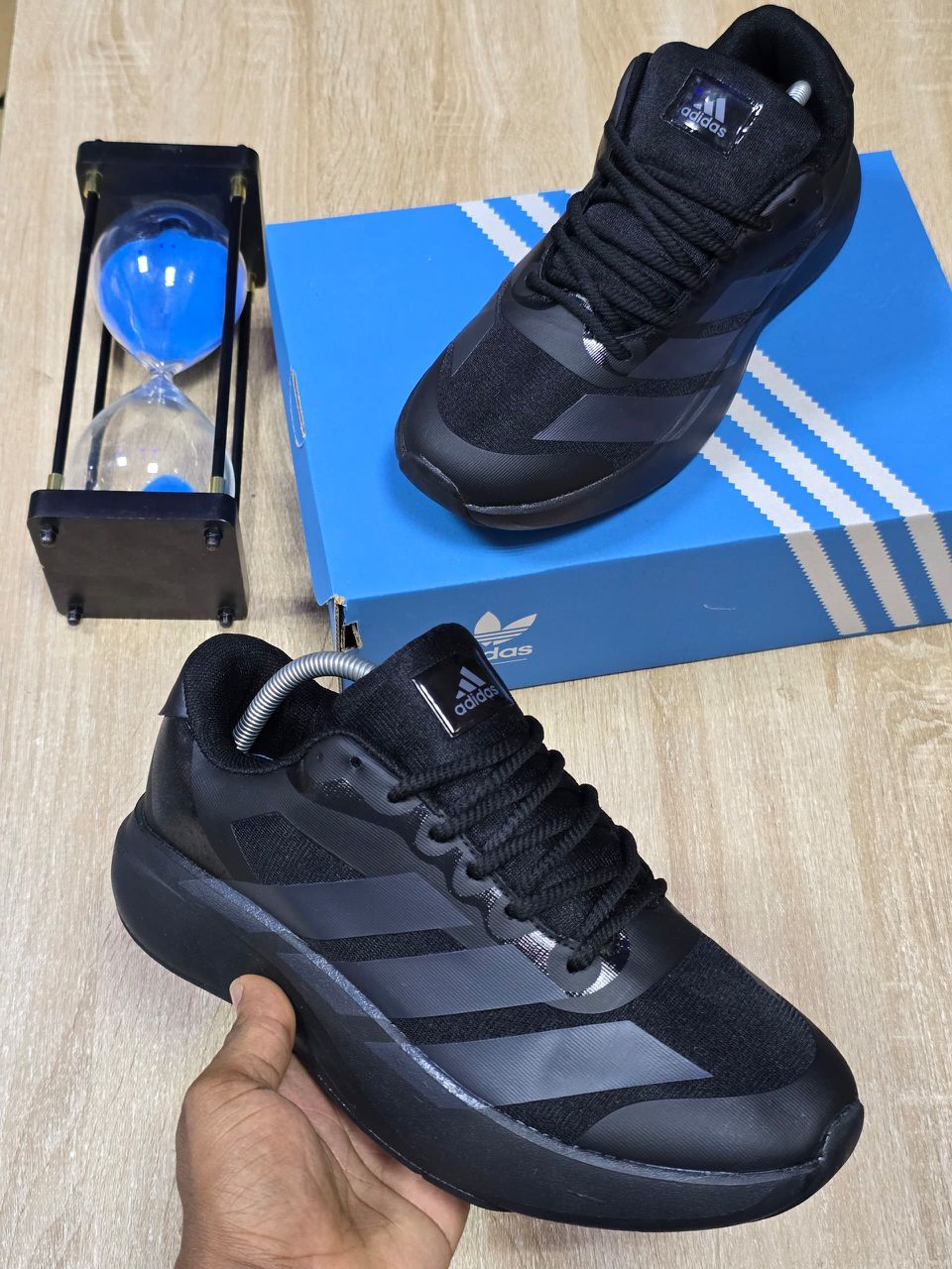Adidas Men’s Luxury Comfort Sneakers A pair of men's luxury black comfort sneakers with grey accents, displayed on a blue box with a hand holding one shoe, showcasing their bold design and thick, cushioned sole.