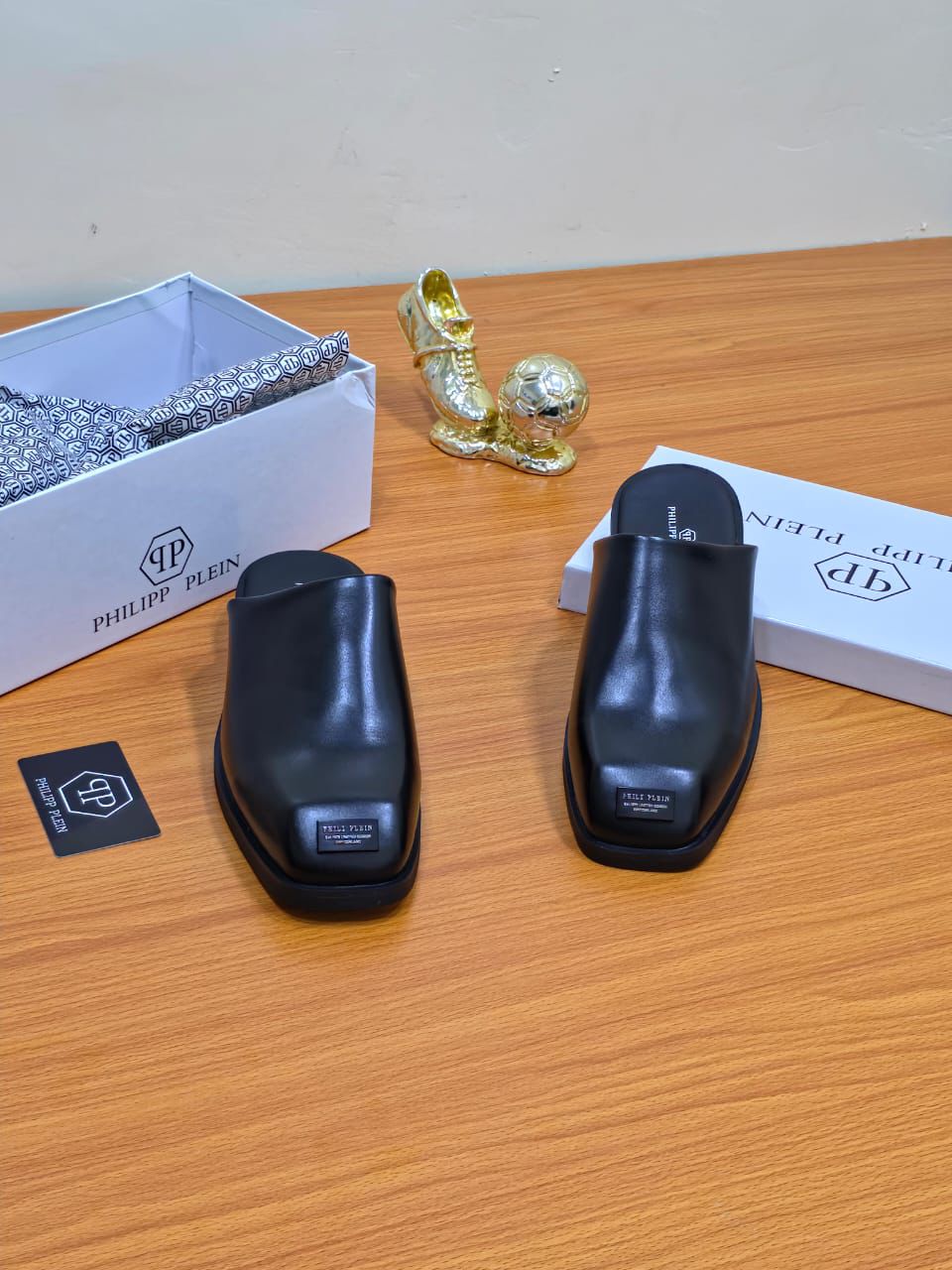 Philipp Plein Men’s Leather Mules A pair of men's luxury black leather mules with a square toe, displayed on a wooden surface next to premium packaging, showcasing their sleek design and premium material.