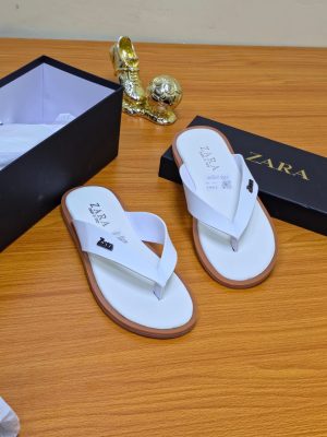 A pair of men's luxury white leather sandals with a brown sole, displayed on a wooden surface next to a black shoebox, showcasing their premium material and elegant design.