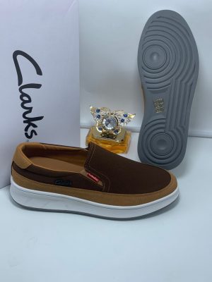 Black and tan Clarks suede slip-on sneakers with white midsole and grey tread displayed with Clarks box and perfume.