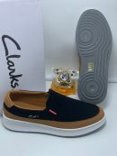 Black and tan Clarks suede slip-on sneakers with white midsole and grey tread displayed with Clarks box and perfume.
