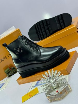 Black leather combat boots with chunky platform sole, lace-up front, and silver shield hardware, displayed beside orange Louis Vuitton box and decorative perfume bottle.