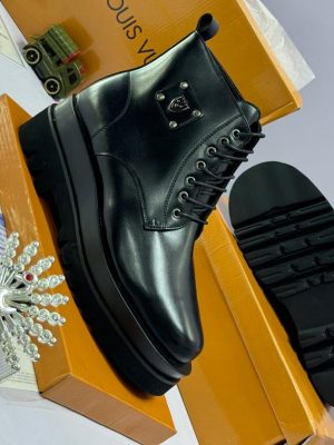 Black leather combat boots with chunky platform sole, lace-up front, and silver shield hardware, displayed beside orange Louis Vuitton box and decorative perfume bottle.