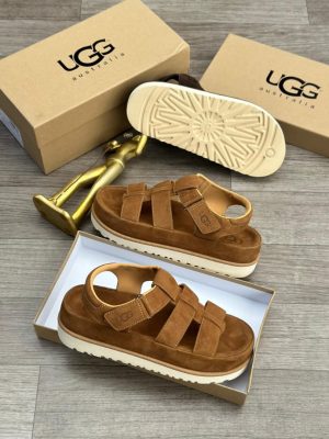 UGG Australia men's brown suede fisherman sandals with thick white sunburst sole, displayed with branded cardboard boxes and gold decorative figure on wooden floor.