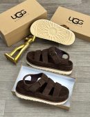 UGG Australia men's brown suede fisherman sandals with thick white sunburst sole, displayed with branded cardboard boxes and gold decorative figure on wooden floor.
