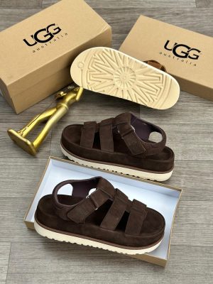 UGG Australia men's brown suede fisherman sandals with thick white sunburst sole, displayed with branded cardboard boxes and gold decorative figure on wooden floor.