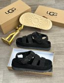 UGG Australia men's brown suede fisherman sandals with thick white sunburst sole, displayed with branded cardboard boxes and gold decorative figure on wooden floor.