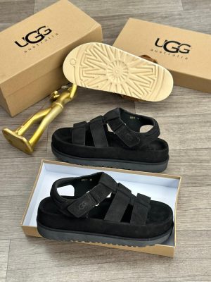 UGG Australia men's brown suede fisherman sandals with thick white sunburst sole, displayed with branded cardboard boxes and gold decorative figure on wooden floor.
