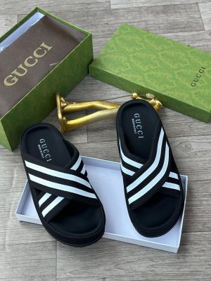 Gucci men's black and white Web stripe slide sandals with rubber footbed, displayed with green Gucci box and gold decorative item on wooden floor.