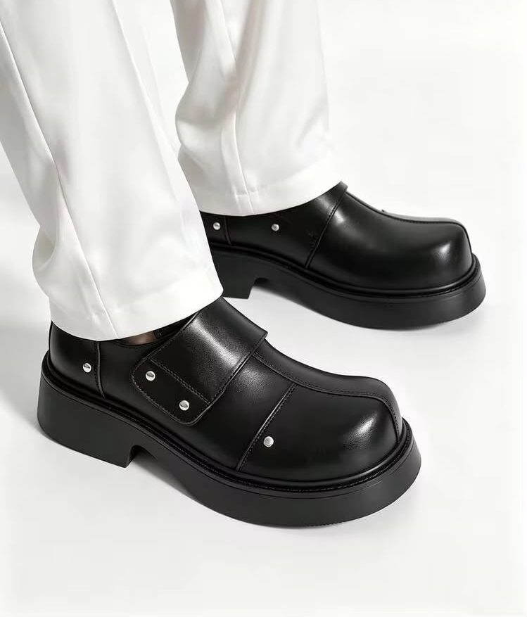 Premium black leather monk-strap loafers Men's luxury black leather monk-strap loafers with chunky black platform soles and silver hardware accents being held by hand next to white trousers.