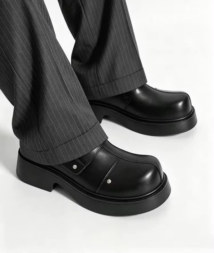 Premium black leather monk-strap loafers Men's luxury black leather monk-strap loafers with chunky black platform soles and silver hardware accents being held by hand next to white trousers.