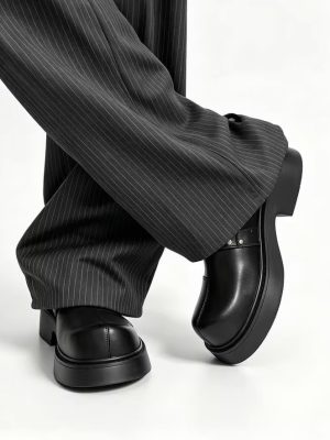 Men's luxury black leather monk-strap loafers with chunky black platform soles and silver hardware accents being held by hand next to white trousers.