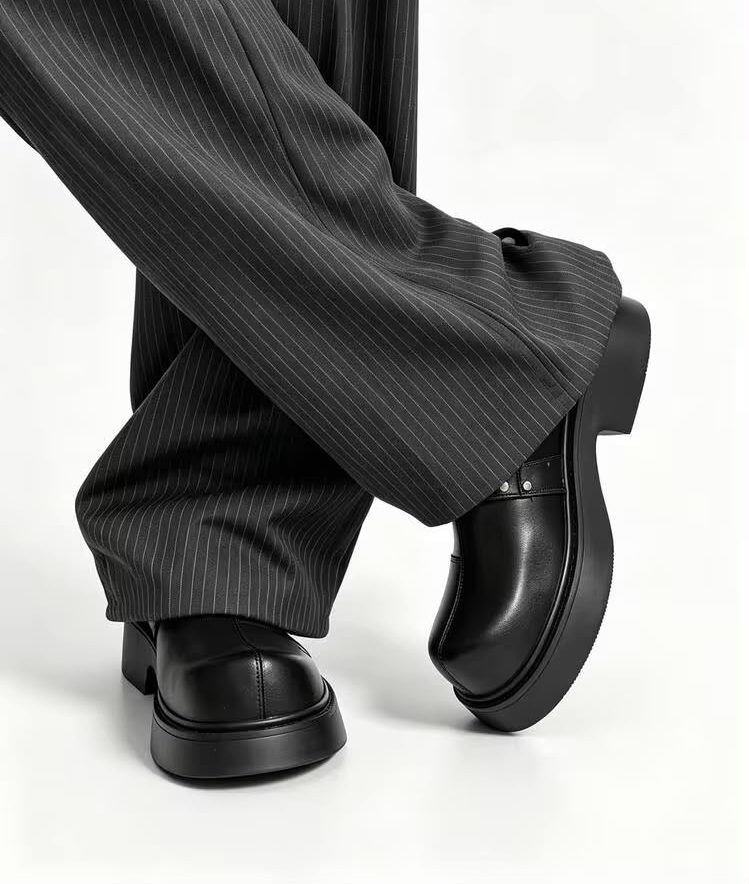 Premium black leather monk-strap loafers Men's luxury black leather monk-strap loafers with chunky black platform soles and silver hardware accents being held by hand next to white trousers.