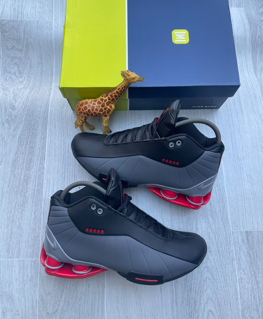 Premium Nike Shox TL sneakers Nike Shox TL men's sneakers in black, anthracite, and red with iconic Shox heel columns, displayed on a wooden floor with a blue and yellow Nike Shox box.