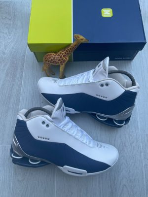 Nike Shox TL men's sneakers in black, anthracite, and red with iconic Shox heel columns, displayed on a wooden floor with a blue and yellow Nike Shox box.