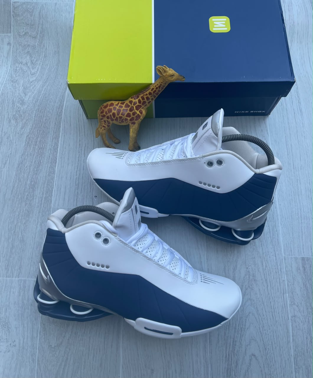 Premium Nike Shox TL sneakers Nike Shox TL men's sneakers in black, anthracite, and red with iconic Shox heel columns, displayed on a wooden floor with a blue and yellow Nike Shox box.