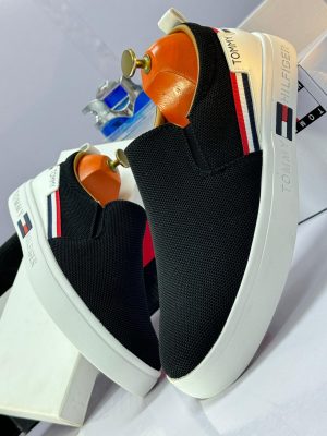 Alt Text: Black knit Tommy Hilfiger slip-on sneakers with white platform sole and red, white, blue flag stripe - luxury men's casual shoes.