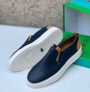 Navy blue Lacoste leather slip-on sneakers with white sole and tan heel accents displayed with green Lacoste box.