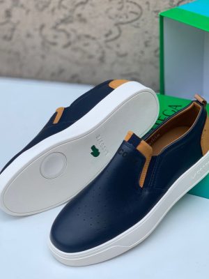 Navy blue Lacoste leather slip-on sneakers with white sole and tan heel accents displayed with green Lacoste box.