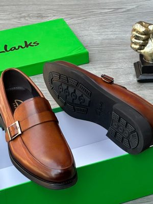 Clarks men’s brown leather monk strap loafers with burnished gradient finish, single buckle detail, and cushioned sole, displayed beside green branded box and golden thumbs-up sculpture on grey wood surface.