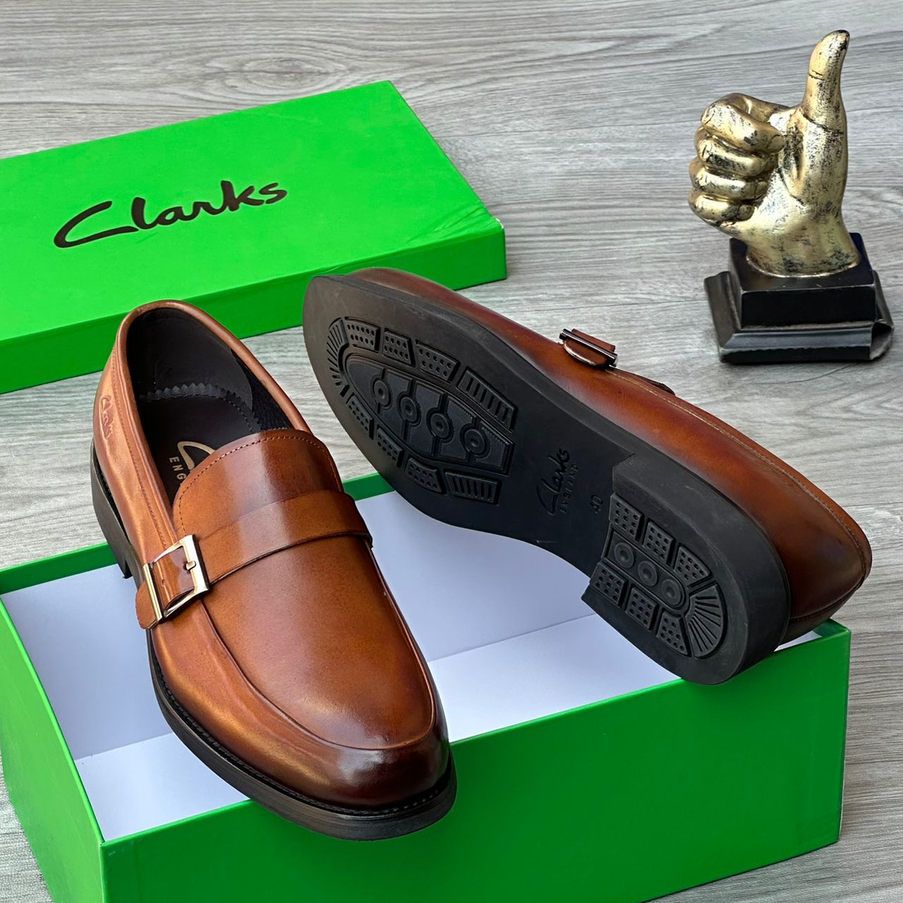 Clarks men’s leather monk strap loafers Clarks men’s brown leather monk strap loafers with burnished gradient finish, single buckle detail, and cushioned sole, displayed beside green branded box and golden thumbs-up sculpture on grey wood surface.