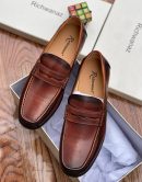 Richwanaz men’s brown leather penny loafers with burnished finish and hand-stitched toe, displayed beside branded box on wooden surface.