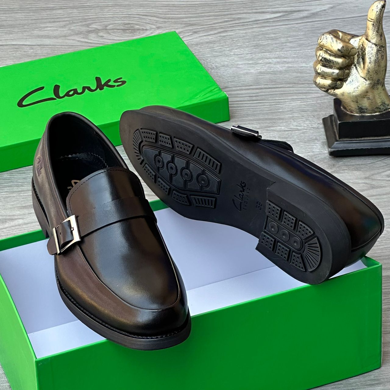 Clarks men’s leather monk strap loafers Clarks men’s brown leather monk strap loafers with burnished gradient finish, single buckle detail, and cushioned sole, displayed beside green branded box and golden thumbs-up sculpture on grey wood surface.
