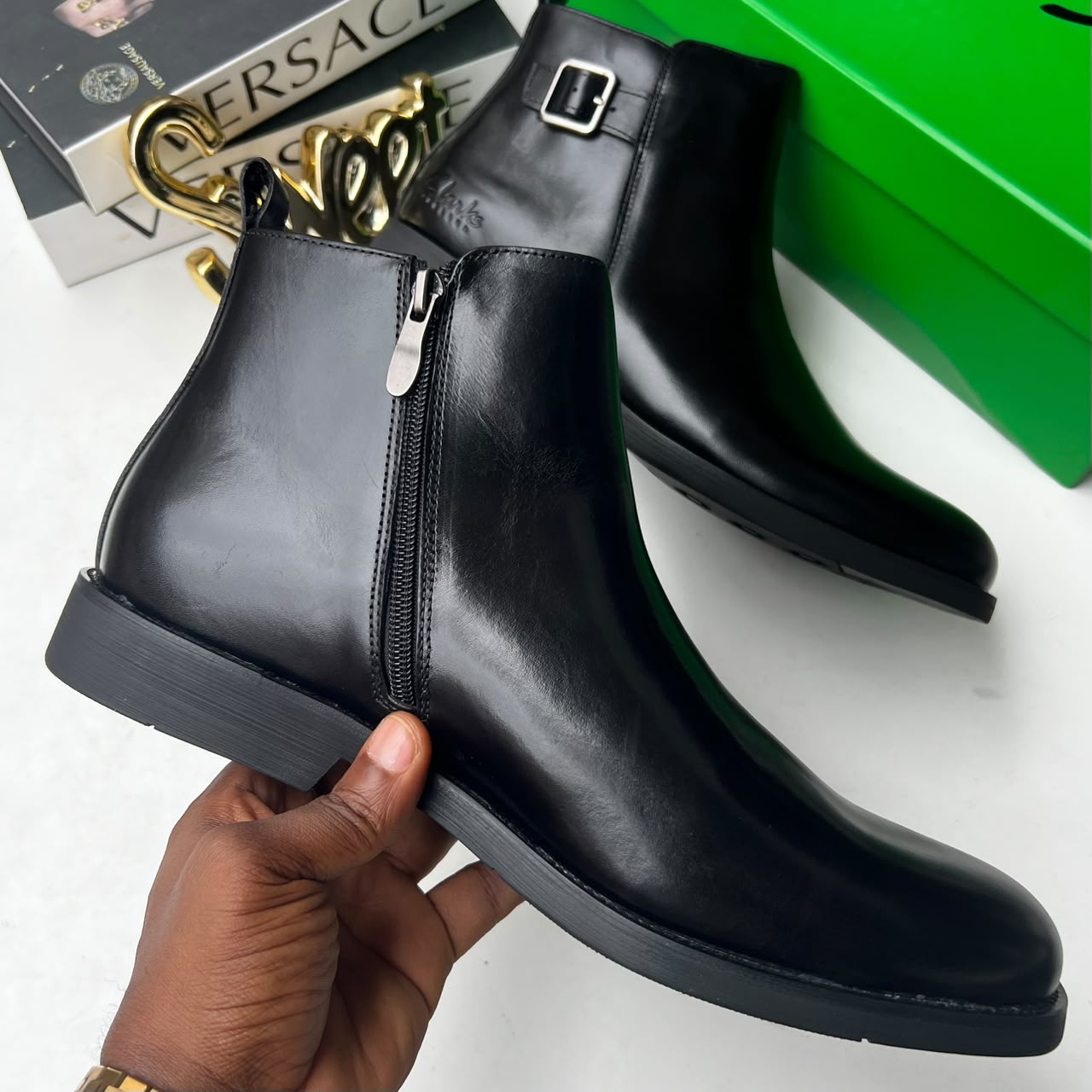 Clarks dark brown leather Chelsea boots Clarks men's dark brown leather Chelsea boots with side buckle strap and black rubber sole, held in hand with branded green shoebox and Versace books in background.