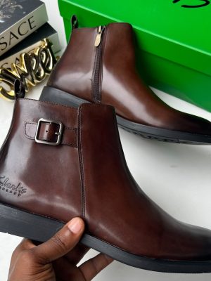 Clarks men's dark brown leather Chelsea boots with side buckle strap and black rubber sole, held in hand with branded green shoebox and Versace books in background.
