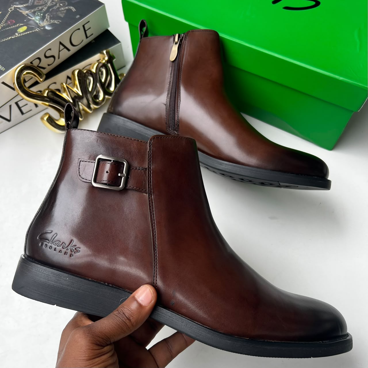 Clarks dark brown leather Chelsea boots Clarks men's dark brown leather Chelsea boots with side buckle strap and black rubber sole, held in hand with branded green shoebox and Versace books in background.