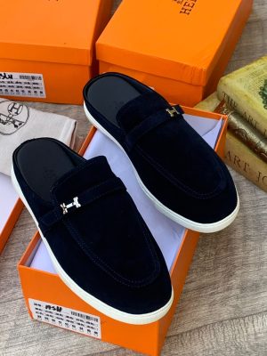 Hermès men's camel suede loafer mules with silver H-hardware and white rubber sole, displayed with orange Hermès box and dust bag on wooden surface.