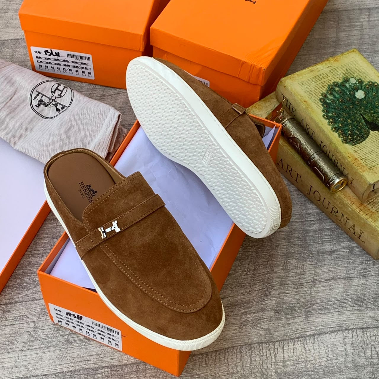 Hermès camel suede loafer mules Hermès men's camel suede loafer mules with silver H-hardware and white rubber sole, displayed with orange Hermès box and dust bag on wooden surface.