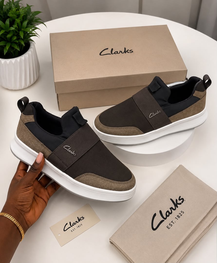 Clarks suede slip-on sneakers Clarks men's olive suede slip-on sneakers with elastic side panels and white rubber sole, held in hand with branded beige shoebox and dust bag on white table.