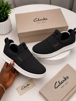 Clarks men's olive suede slip-on sneakers with elastic side panels and white rubber sole, held in hand with branded beige shoebox and dust bag on white table.