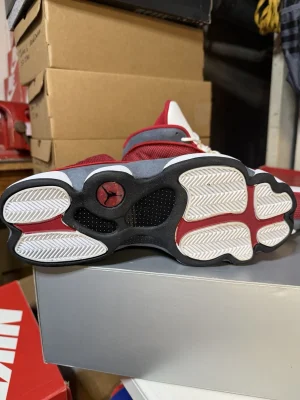 White, black, and grey Air Jordan 13 high-top sneakers with holographic eye detail and paw print outsole displayed on black box.