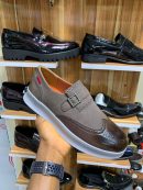 Hand holding a brown and taupe suede leather hybrid loafer with a metal buckle and thick white sole, displayed in front of a shelf of patent leather shoes.