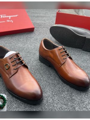 Salvatore Ferragamo men’s cognac brown leather oxford shoes with brogue detailing and Gancini hardware, displayed beside red branded box on grey carpet background.