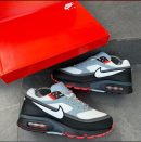Nike Air Max 90 men’s sneakers in grey, white, black, and red with visible Air unit and waffle sole, displayed beside red branded box on dark tiled surface.