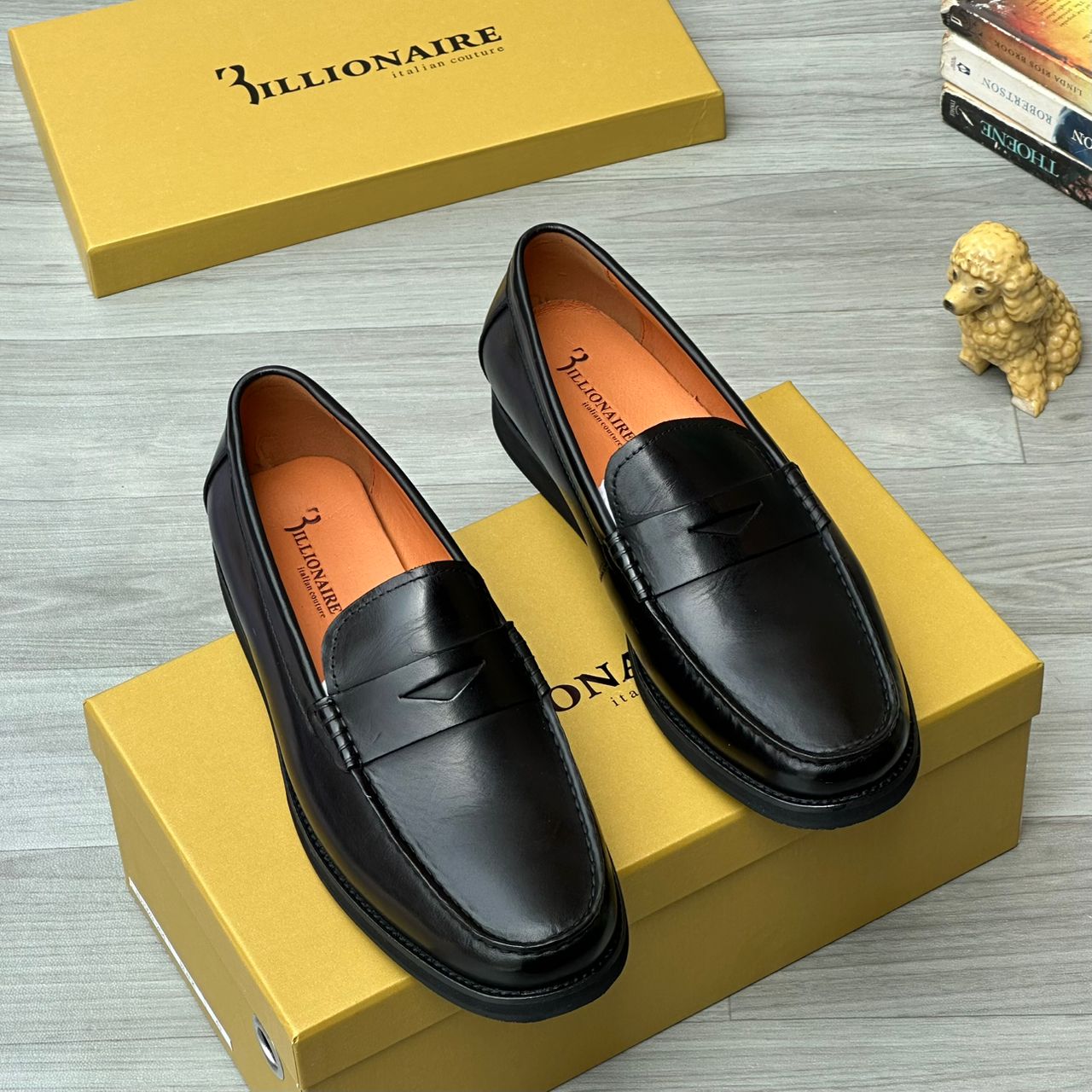 Italian opulence Billionaire men’s penny loafers Billionaire Italian Couture men’s brown burnished leather penny loafers with moccasin stitching and rugged lug sole, displayed beside mustard-yellow branded box, poodle figurine, and stacked books on light wood surface.