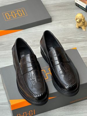 Oggi Shoes men’s brown leather penny loafers with hand-etched script design, burnished gradient finish, and rugged lug sole, displayed beside grey-and-orange branded box, poodle figurine, and stacked books on light wood surface.