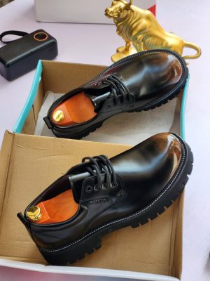 Men's luxury black leather derby boots with burnished brown toe cap, thick black lug sole, and contrast stitching, displayed next to a gold bull statue and colorful shoebox.