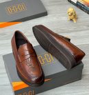 Oggi Shoes men’s brown leather penny loafers with hand-etched script design, burnished gradient finish, and rugged lug sole, displayed beside grey-and-orange branded box, poodle figurine, and stacked books on light wood surface.